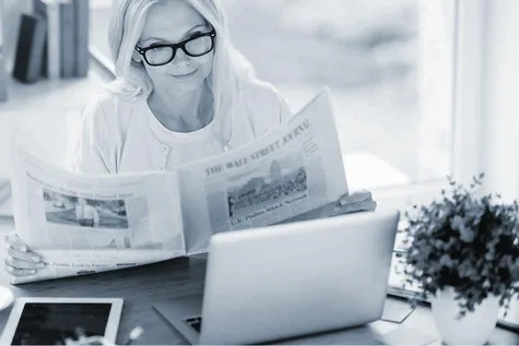 Eine Frau, die Zeitung liest. Vor ihr auf dem Tisch liegen ein Laptop und ein Tablet